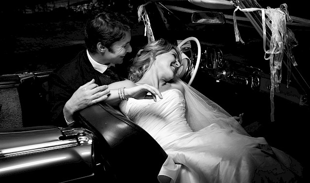 Bride leaning back into her husband, sharing a laugh in an open-topped sports car – photo by Greg Lumley