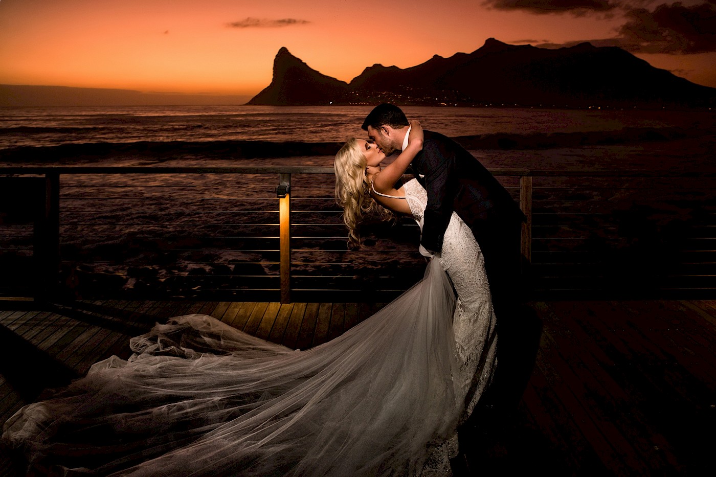 A groom kisses his beautiful bride as the sun sets, with the ocean and a majestic mountain as their backdrop. Photo by Greg Lumley.